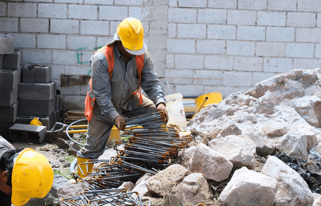 Construction workers in safety gear are moving and sorting steel bars and rocks. Latent Conditions