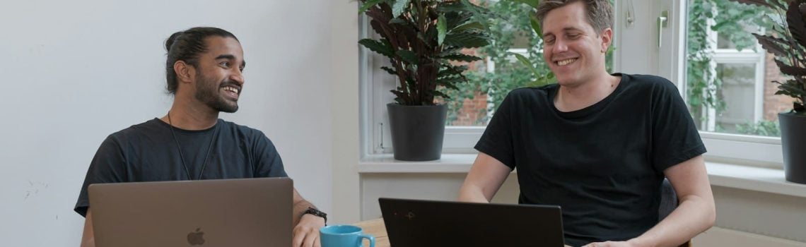 Two men sitting at a brown table with laptops. Startup Funding Landscape. Small Business Management Consulting. Business Growth Consultant. Business Growth Strategist.
